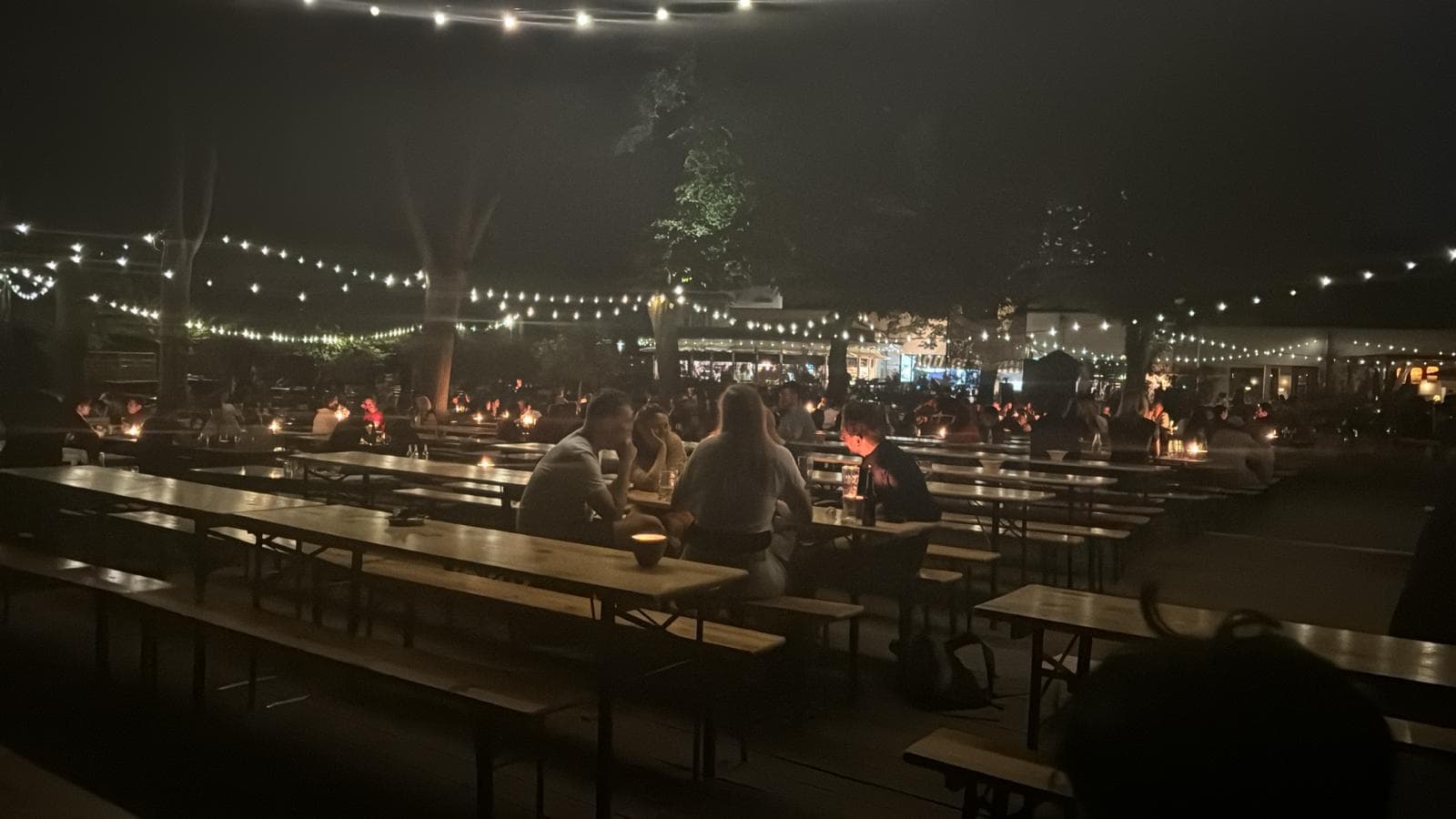 Cozy beer garden at night with string lights and people chatting.